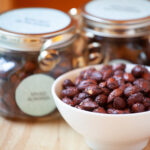 A white ceramic bowl contains spiced almonds. Decorative jars with silver ribbon sit beside the bowl, and a wooden table.