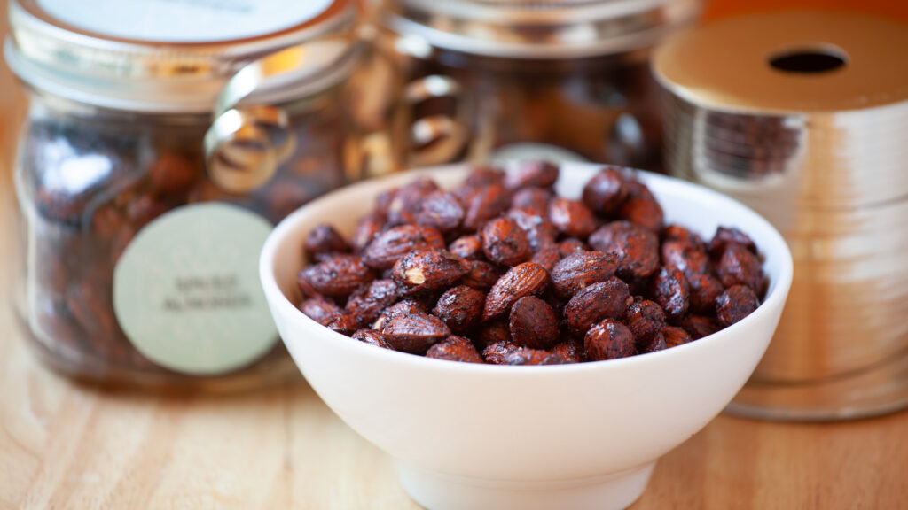 A white ceramic bowl contains holiday spiced nuts. Decorative jars with silver ribbon sit beside the bowl, and a wooden table.