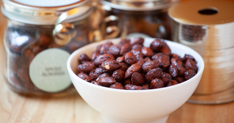 A white ceramic bowl contains spiced almonds. Decorative jars with silver ribbon sit beside the bowl, and a wooden table.