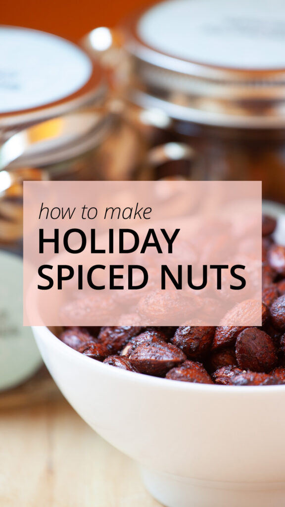 A white ceramic bowl containing holiday spiced nuts sit beside the bowl, and a wooden table.