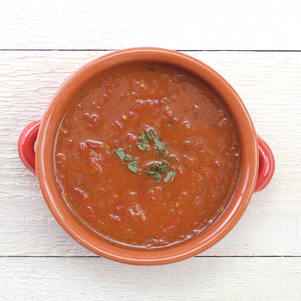 An overhead view of a red bowl with roasted tomato soup, garnished with thyme leaves, on a background of white wooden planks.