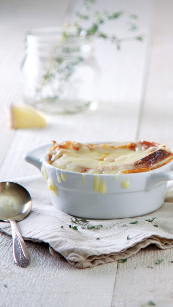 A white bowl is filled with homemade roasted tomato soup, topped with a slice of rustic bread, and gooey, melted gruyere cheese. It sits on a beige napkin along with a silver soup spoon.
