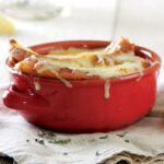 A red bowl is filled with homemade roasted tomato soup, topped with a slice of rustic bread, and gooey, melted gruyere cheese. It sits on a beige napkin along with a silver soup spoon and another chunk of crusty bread.