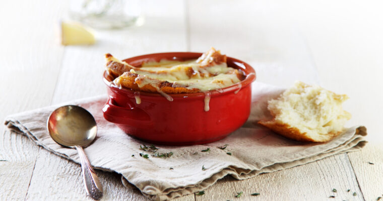 A red bowl is filled with homemade roasted tomato soup, topped with a slice of rustic bread, and gooey, melted gruyere cheese. It sits on a beige napkin along with a silver soup spoon and another chunk of crusty bread.