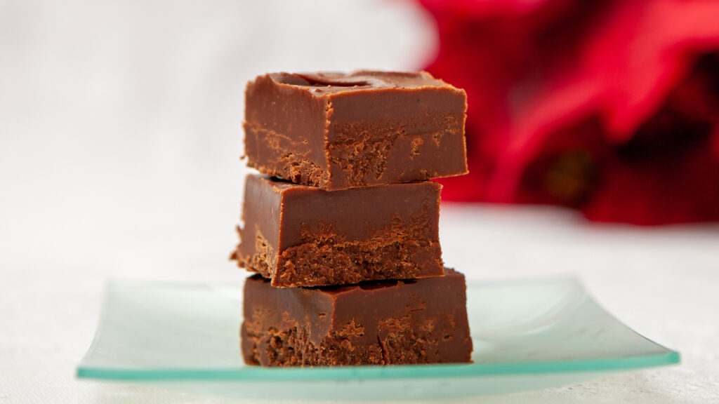Three pieces of chocolate fudge are stacked on a glass plate, on a table with a white tablecloth. In the background is a red poinsettia plant.