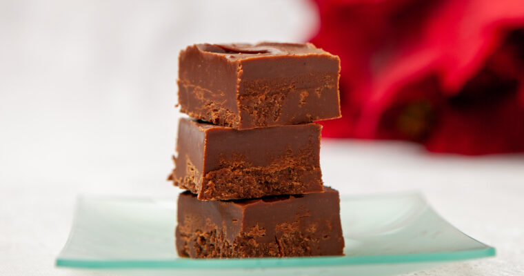 Three pieces of chocolate fudge are stacked on a glass plate, on a table with a white tablecloth. In the background is a red poinsettia plant.