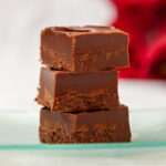 Three pieces of chocolate fudge are stacked on a glass plate, on a table with a white tablecloth. In the background is a red poinsettia plant.
