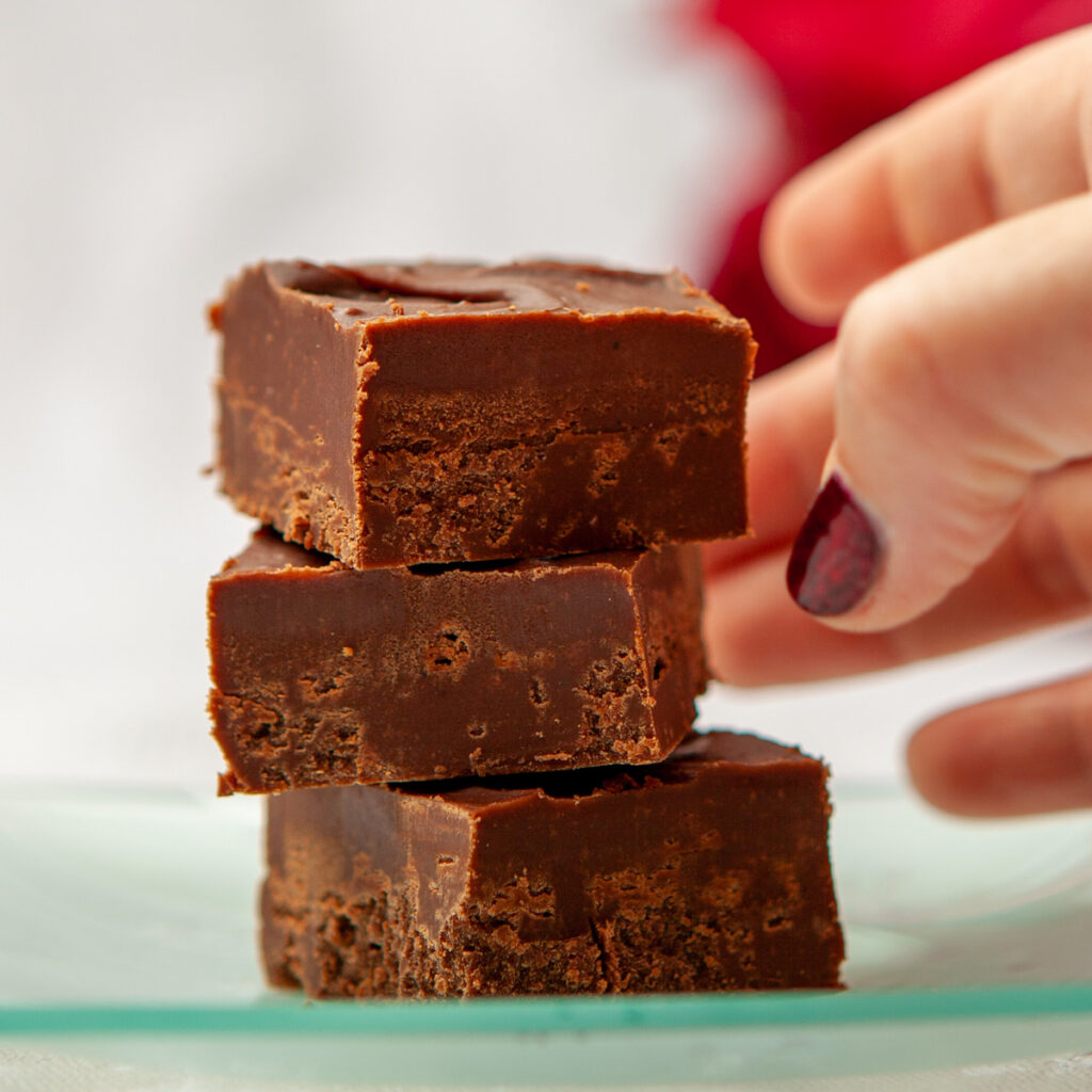 Three pieces of chocolate fudge are stacked on a glass plate. A hand with red sparkly nail polish is reaching for the top piece. The plate is on a table with a white tablecloth. In the background, out of focus, is a red poinsettia plant.