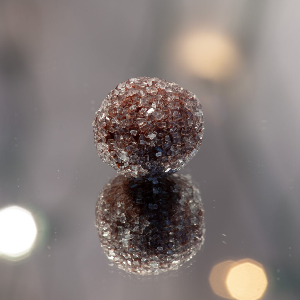 A single chocolate rum ball covered in sparkly sugar sits on a mirrored surface.