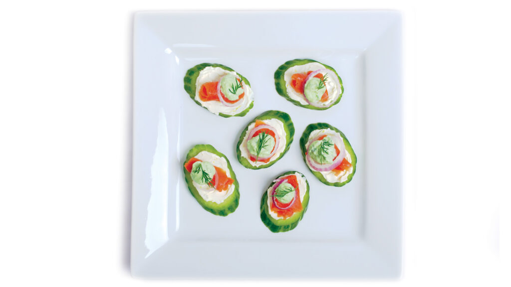 Pieces of thinly sliced smoked salmon lox sit on a smear of lemon cream cheese, topped with a tiny ring of red onion, a dollop of bright green chive cream, and a sprig of fresh dill. Chopped chives are sprinkled on the plate.