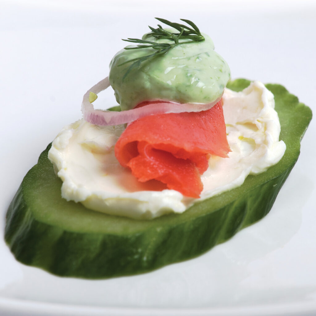 Pieces of thinly sliced smoked salmon lox sit on a smear of lemon cream cheese, topped with a tiny ring of red onion, a dollop of bright green chive cream, and a sprig of fresh dill. Chopped chives are sprinkled on the plate.