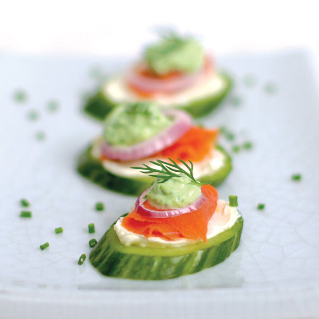 Pieces of thinly sliced smoked salmon lox sit on a smear of lemon cream cheese, topped with a tiny ring of red onion, a dollop of bright green chive cream, and a sprig of fresh dill. Chopped chives are sprinkled on the plate.