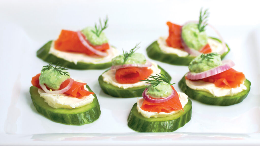 Pieces of thinly sliced smoked salmon lox sit on a smear of lemon cream cheese, topped with a tiny ring of red onion, a dollop of bright green chive cream, and a sprig of fresh dill. Chopped chives are sprinkled on the plate.