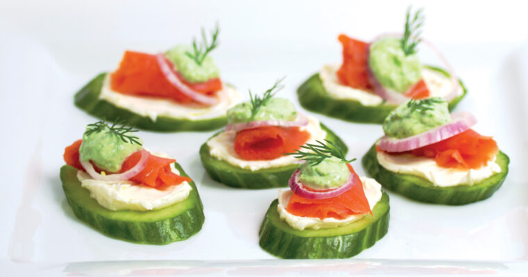 Pieces of thinly sliced smoked salmon lox sit on a smear of lemon cream cheese, topped with a tiny ring of red onion, a dollop of bright green chive cream, and a sprig of fresh dill. Chopped chives are sprinkled on the plate.