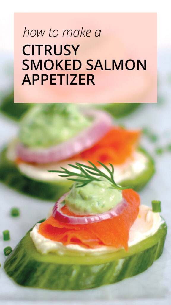 Pieces of thinly sliced smoked salmon lox sit on a smear of lemon cream cheese, topped with a tiny ring of red onion, a dollop of bright green chive cream, and a sprig of fresh dill. Chopped chives are sprinkled on the plate.