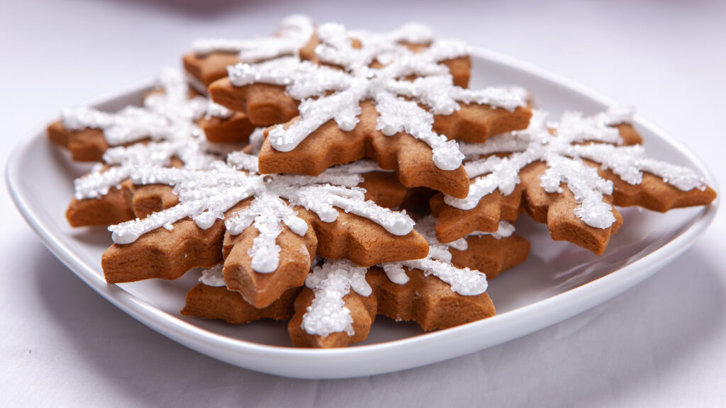 Snowflake shaped gingerbread cookies sit on a white plate. The cookies are decorated with white sparkle sugar and silver decorations.