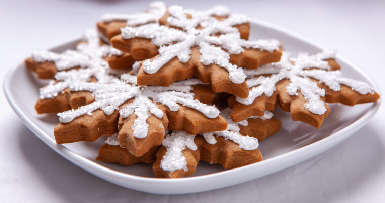 Snowflake shaped gingerbread cookies sit on a white plate. The cookies are decorated with white sparkle sugar and silver decorations.