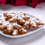 Snowflake shaped gingerbread cookies sit on a white plate. The cookies are decorated with white sparkle sugar and silver decorations.