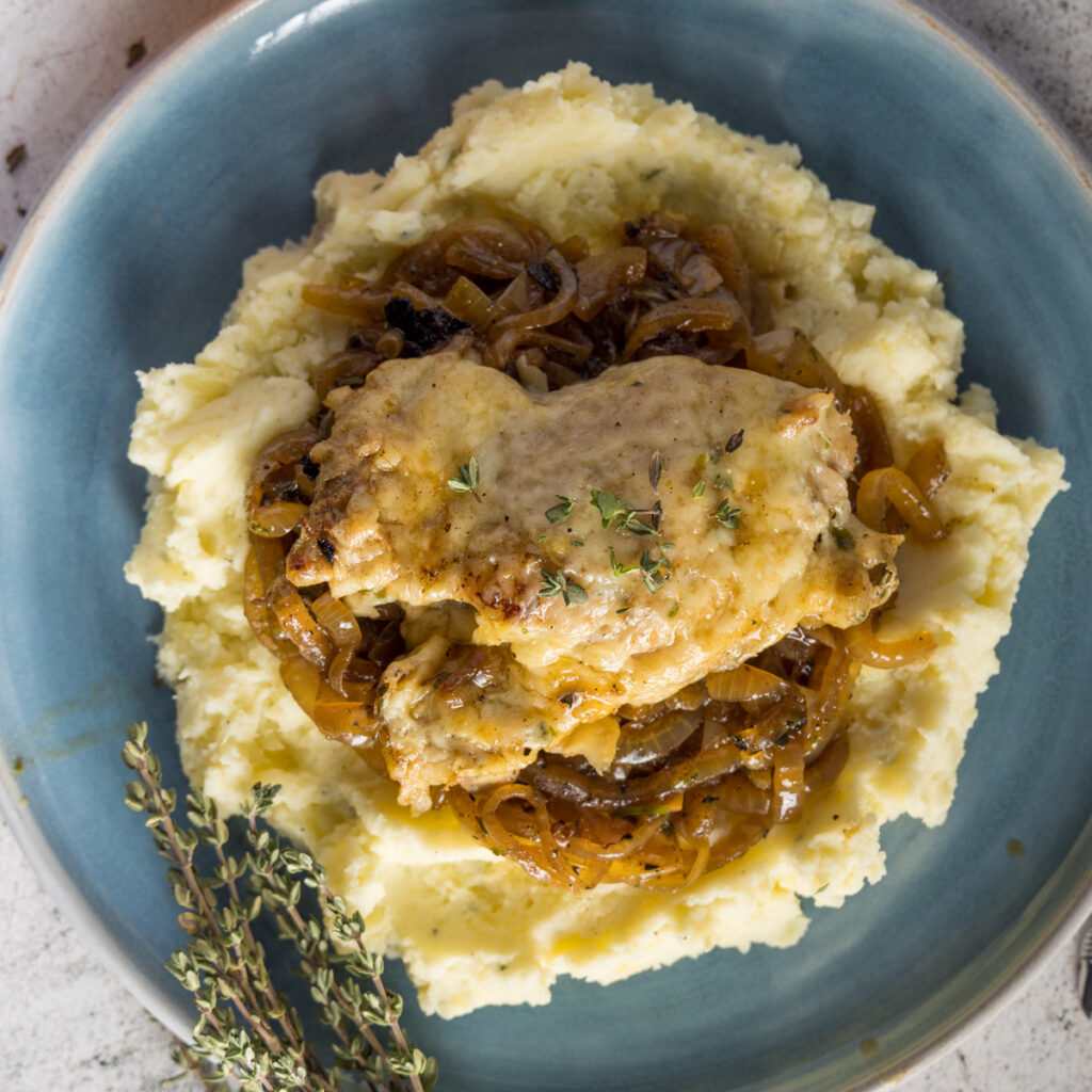 Chicken thighs, topped with melted Gruyère cheese, on top of caramelized onions, mashed potatoes, on a blue plate. Next to the plate is an onion and bunches of fresh thyme leaves.