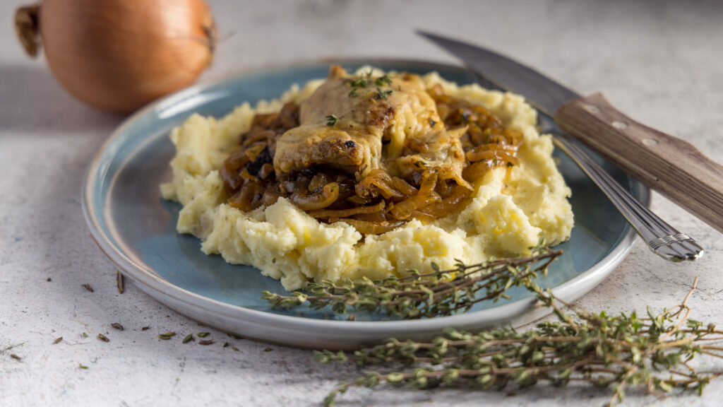 Chicken thighs, topped with melted Gruyère cheese, on top of caramelized onions, mashed potatoes, on a blue plate. Next to the plate is an onion and bunches of fresh thyme leaves.