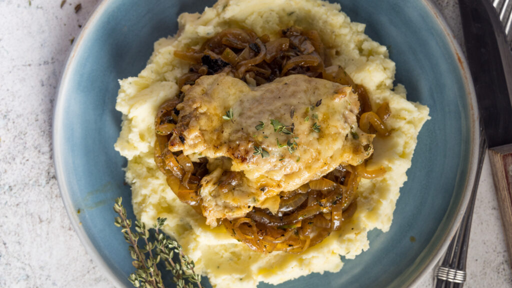 Chicken thighs, topped with melted Gruyère cheese, on top of caramelized onions, mashed potatoes, on a blue plate. Next to the plate is an onion and bunches of fresh thyme leaves.