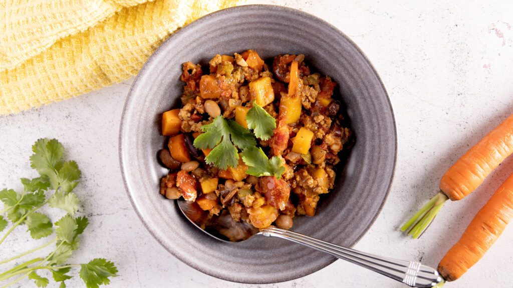 A bowl of turkey chili. In the chili are carrots, squash, beans, and tomatoes. The chili is garnished with cilantro leaves. A spoon rests in the bowl. Next the bowl is a yellow towel, more cilantro, and 2 unpeeled carrots.