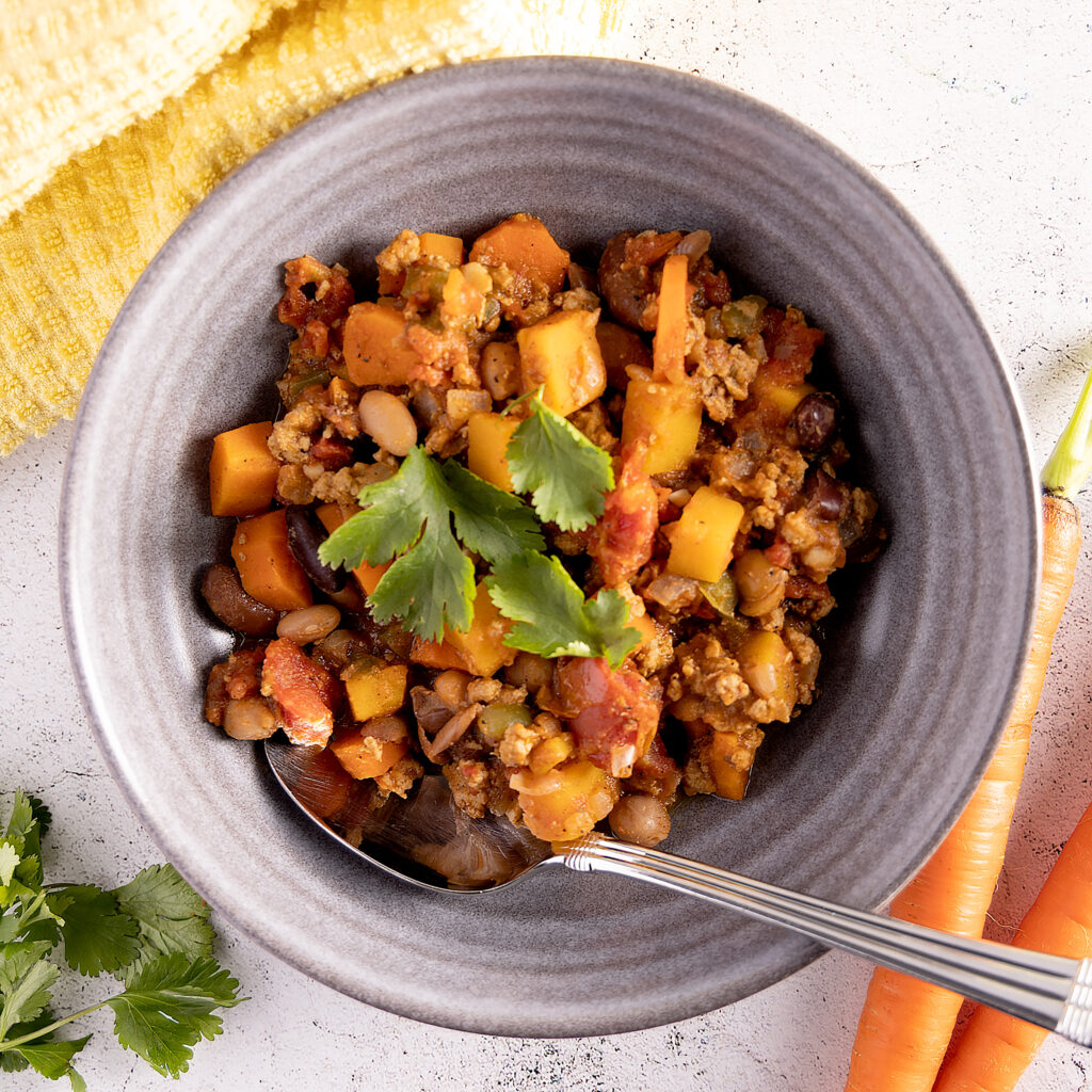 A bowl of turkey chili. In the chili are carrots, squash, beans, and tomatoes. The chili is garnished with cilantro leaves. A spoon rests in the bowl. Next the bowl is a yellow towel, more cilantro, and 2 unpeeled carrots.