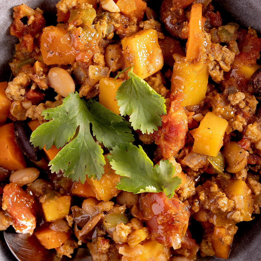 Photo alt:
A bowl of turkey chili. In the chili are carrots, squash, beans, and tomatoes. The chili is garnished with cilantro leaves. A spoon rests in the bowl. Next the bowl is a yellow towel, more cilantro, and 2 unpeeled carrots.
