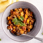 A bowl of turkey chili. In the chili are carrots, squash, beans, and tomatoes. The chili is garnished with cilantro leaves. A spoon rests in the bowl. Next the bowl is a yellow towel, more cilantro, and 2 unpeeled carrots.