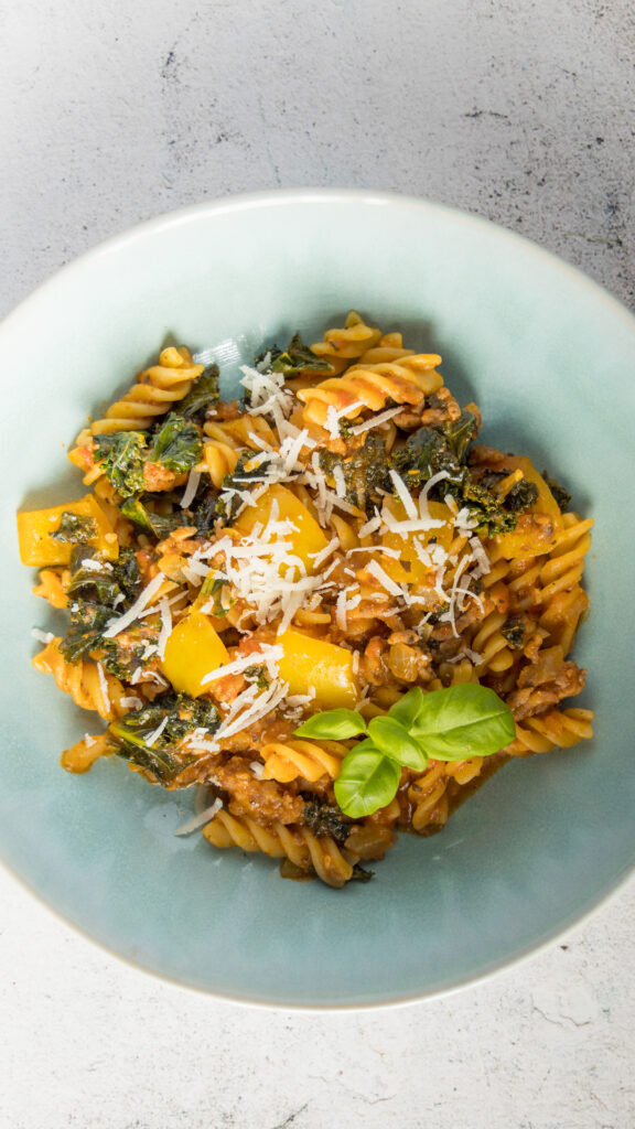 A bowl of sausage pasta with kale, yellow bell peppers, and spices.