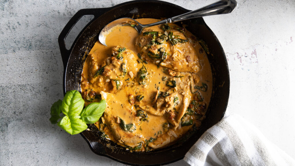 Juicy browned chicken breasts sit in a creamy parmesan sauce, with sundried tomatoes and spinach, in a cast iron pan. A grey and white towel is wrapped around the handle of the pan and a large serving spoon sits in the sauce.