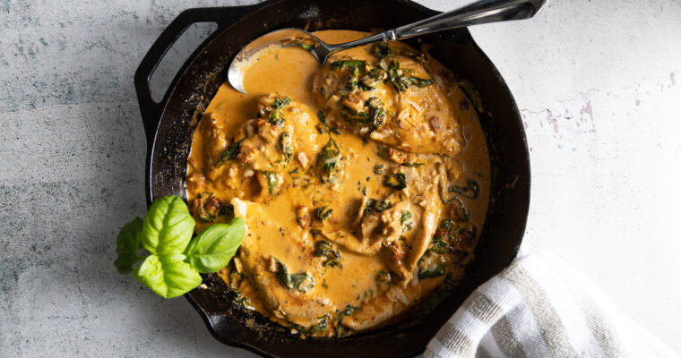 Juicy browned chicken breasts sit in a creamy parmesan sauce, with sundried tomatoes and spinach, in a cast iron pan. A grey and white towel is wrapped around the handle of the pan and a large serving spoon sits in the sauce.