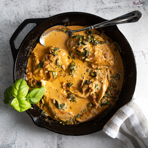 Juicy browned chicken breasts sit in a creamy parmesan sauce, with sundried tomatoes and spinach, in a cast iron pan. A grey and white towel is wrapped around the handle of the pan and a large serving spoon sits in the sauce.