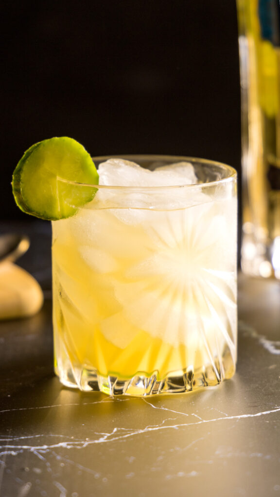 A crystal rocks glass filled with ice holds a pale yellow cocktail, which contains Irish whiskey, elderflower liqueur, lemon juice and cucumber. The glass is garnished with a slice of cucumber. In the background, are a bottle of St-Germain liqueur, Jameson Irish whiskey, and a cutting board with lemon and cucumbers.