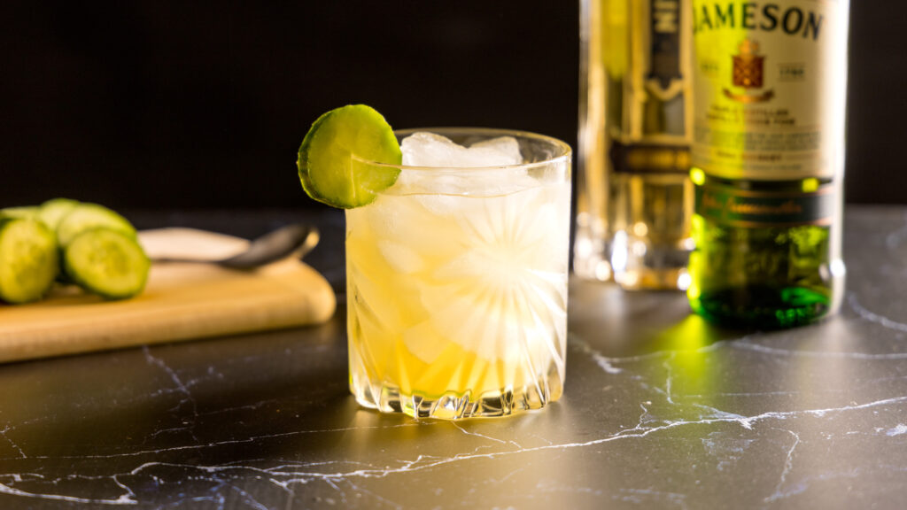 A crystal rocks glass filled with ice holds a pale yellow cocktail, an Irish Maid, which contains Irish whiskey, elderflower liqueur, lemon juice and cucumber. The glass is garnished with a slice of cucumber. In the background, are a bottle of St-Germain liqueur, Jameson Irish whiskey, and a cutting board with lemon and cucumbers.