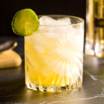 A crystal rocks glass filled with ice holds a pale yellow cocktail, an Irish Maid, which contains Irish whiskey, elderflower liqueur, lemon juice and cucumber. The glass is garnished with a slice of cucumber. In the background, are a bottle of St-Germain liqueur, Jameson Irish whiskey, and a cutting board with lemon and cucumbers.