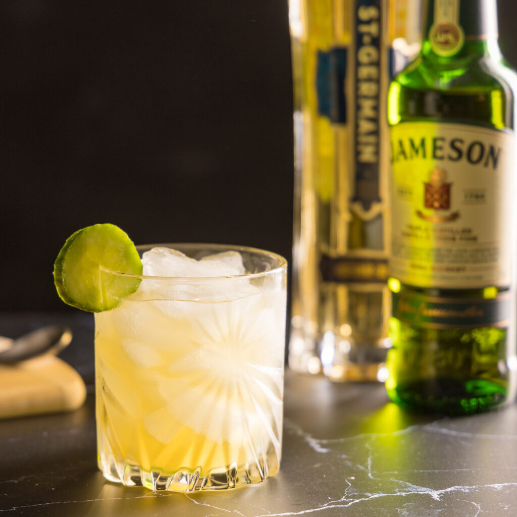 A crystal rocks glass filled with ice holds a pale yellow cocktail, an Irish Maid, which contains Irish whiskey, elderflower liqueur, lemon juice and cucumber. The glass is garnished with a slice of cucumber. In the background, are a bottle of St-Germain liqueur, Jameson Irish whiskey, and a cutting board with lemon and cucumbers.