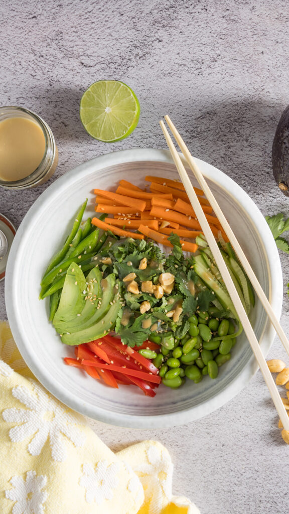 In bowl are rice noodles, sliced vegetables & herbs, sprinkled with sesame seeds and dressing. Next to the bowl are chopsticks, a whole avocado, half a lime, peanuts, bunches of cilantro and mint, and an open jar of dressing with a spoon next to it.