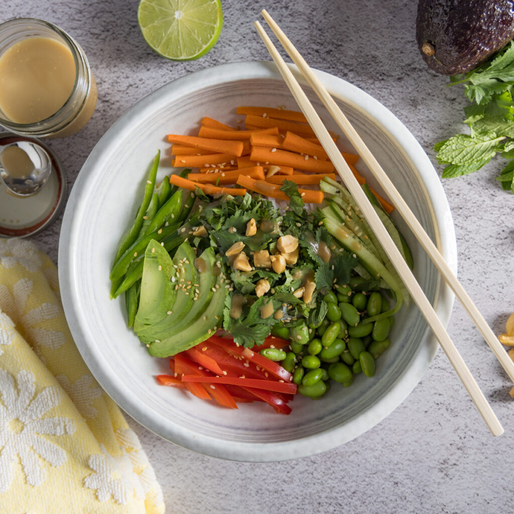 In bowl are rice noodles, sliced vegetables & herbs, sprinkled with sesame seeds and dressing. Next to the bowl are chopsticks, a whole avocado, half a lime, peanuts, bunches of cilantro and mint, and an open jar of dressing with a spoon next to it.