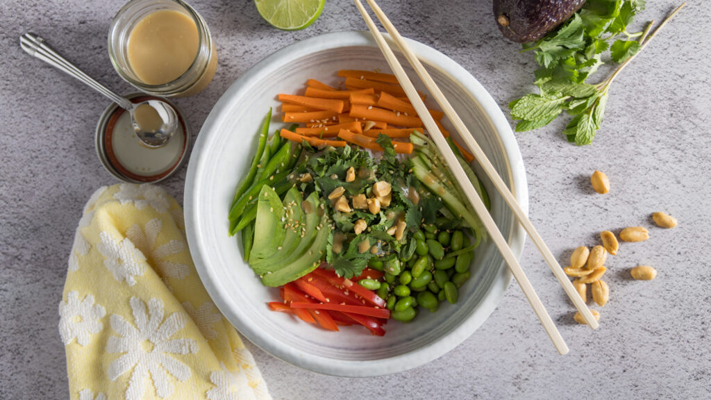 In bowl are rice noodles, sliced vegetables & herbs, sprinkled with sesame seeds and dressing. Next to the bowl are chopsticks, a whole avocado, half a lime, peanuts, bunches of cilantro and mint, and an open jar of dressing with a spoon next to it.