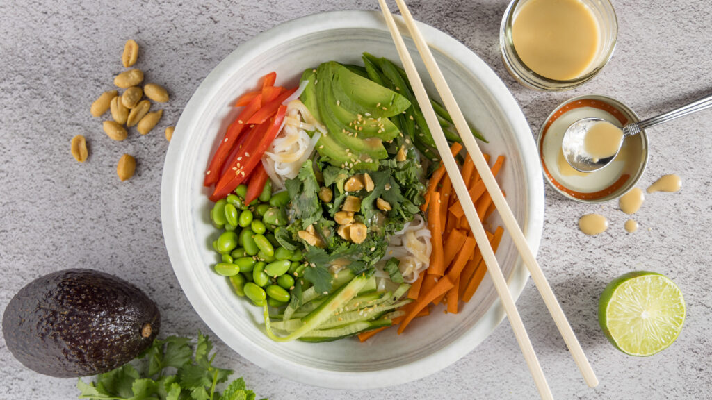 In bowl are rice noodles, sliced vegetables & herbs, sprinkled with sesame seeds and dressing. Next to the bowl are chopsticks, a whole avocado, half a lime, peanuts, bunches of cilantro and mint, and an open jar of dressing with a spoon next to it.