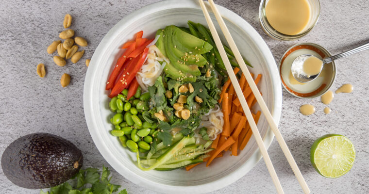 In bowl are rice noodles, sliced vegetables & herbs, sprinkled with sesame seeds and dressing. Next to the bowl are chopsticks, a whole avocado, half a lime, peanuts, bunches of cilantro and mint, and an open jar of dressing with a spoon next to it.