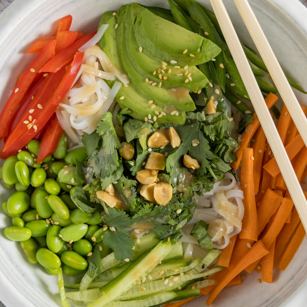 In bowl are rice noodles, sliced vegetables & herbs, sprinkled with sesame seeds and dressing.