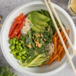 In bowl are rice noodles, sliced vegetables & herbs, sprinkled with sesame seeds and dressing. Next to the bowl are chopsticks, a whole avocado, half a lime, peanuts, bunches of cilantro and mint, and an open jar of dressing with a spoon next to it.