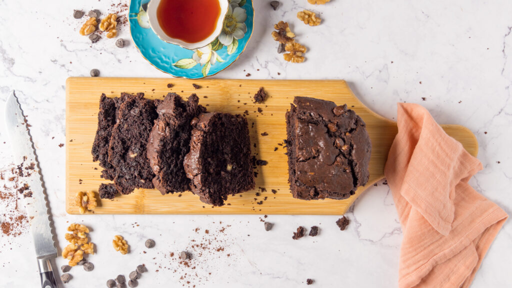 An overhead view of a loaf of chocolate banana bread, on a wooden cutting board, partially sliced up. 