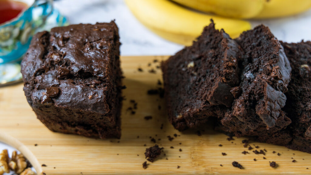 A loaf of chocolate banana bread sits on a wooden cutting board, partially sliced up.