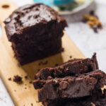 A loaf of chocolate banana bread sits on a wooden cutting board, partially sliced up.