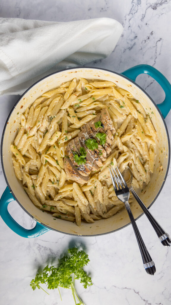 In a large 2-handled skillet, pasta with a creamy sauce, sliced grilled chicken, and garnished with fresh parsley and parmesan cheese.