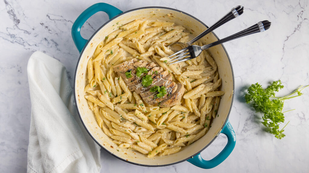 In a large 2-handled skillet, pasta with a creamy sauce, sliced grilled chicken, and garnished with fresh parsley and parmesan cheese.
