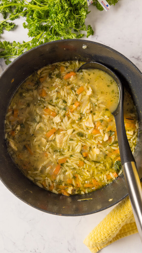 A pot of lemon chicken orzo soup, with a ladle, and a yellow towel.